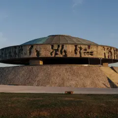 Monumentalne mauzoleum o okrągłej podstawie, z betonowym dyskiem ozdobionym płaskorzeźbami i szklanym dachem.