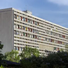 Wielopiętrowy budynek z kolorowymi balkonami, symbol epoki modernizmu, otoczony zielenią.