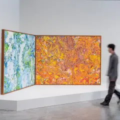 Dwie abstrakcyjne, marmurkowe obrazy w drewnianych ramach, jeden niebiesko-zielony, drugi pomarańczowo-żółty, prezentowane na białej platformie podczas biennale.