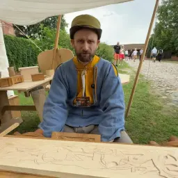 Mistrz snycerstwa prezentuje swoje dzieło. Na stole leży drewniana deska z wyrytymi wzorami, a obok małe drewniane pudełka.