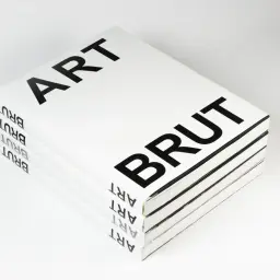 Stos białych książek z czarnym napisem "ART BRUT" na okładce. Na grzbietach powtarza się napis "ART BRUT".
