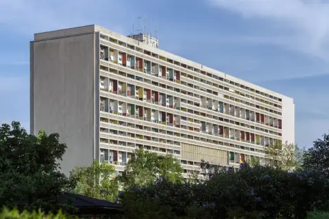 Wielopiętrowy budynek z kolorowymi balkonami, symbol epoki modernizmu, otoczony zielenią.