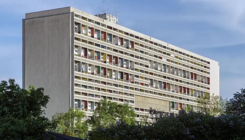 Wielopiętrowy budynek z kolorowymi balkonami, symbol epoki modernizmu, otoczony zielenią.