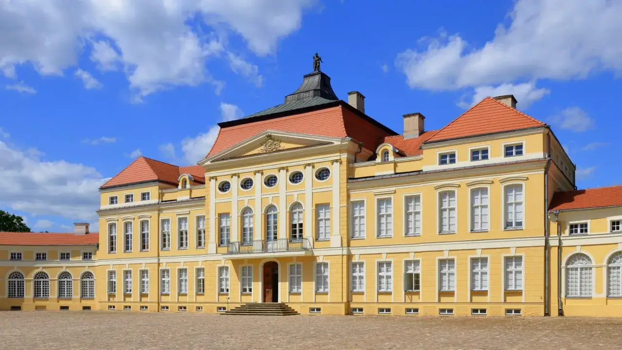 Żółty pałac z czerwonym dachem, symbol oświecenia w Polsce, na tle błękitnego nieba z chmurami.